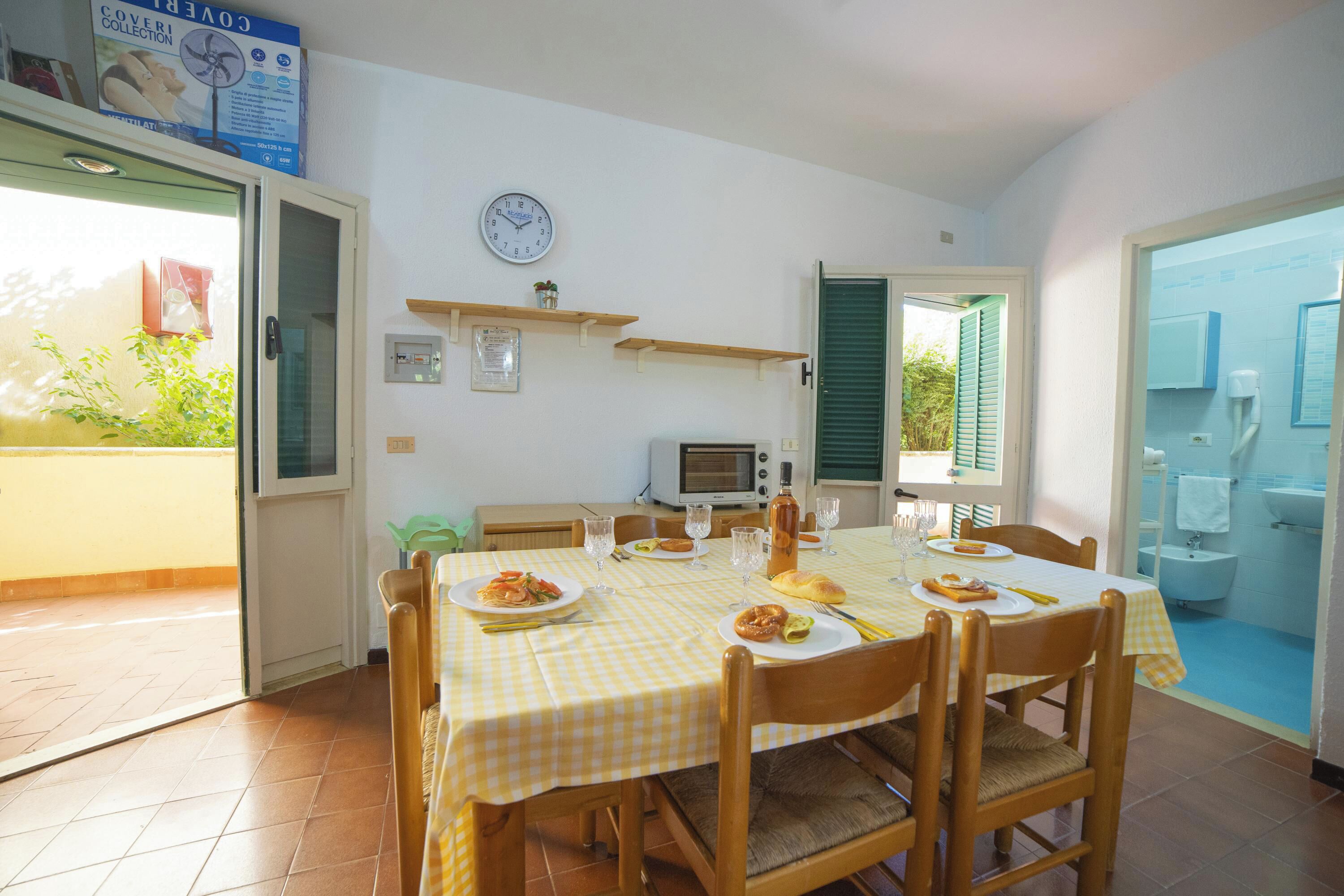 A dining area with a checkered tablecloth, wooden chairs, a clock, and a view of the outside.