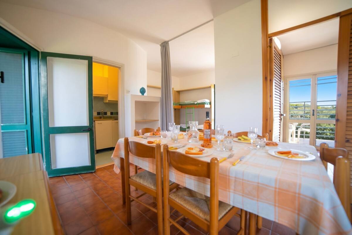 A dining area with a table set for four, a kitchen in the background, and a balcony with a view.