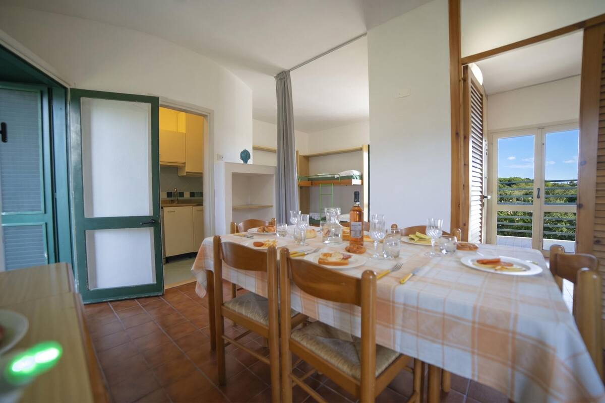 A dining area with a table set for four, chairs, a kitchen in the background, and a view through the window.