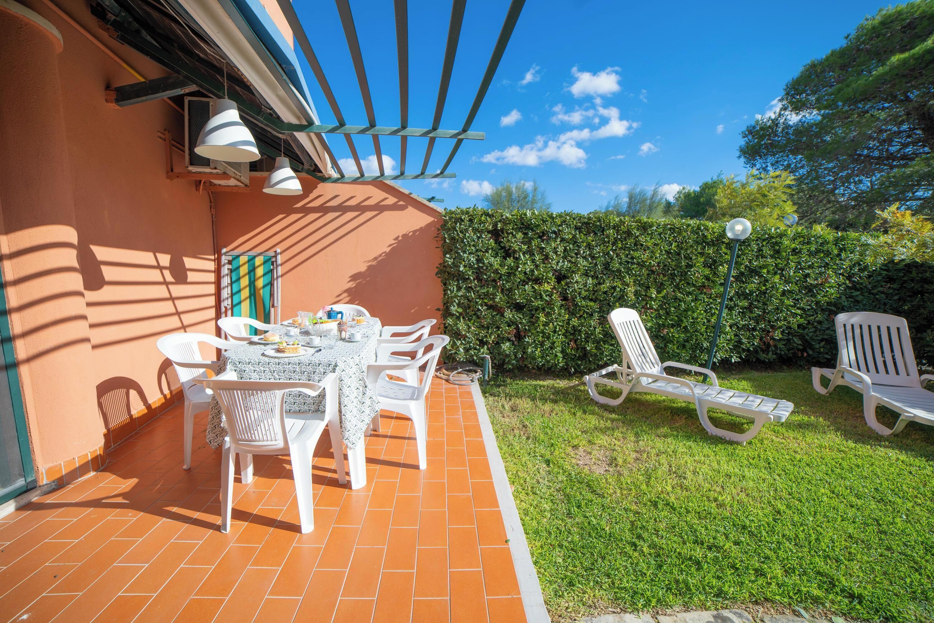 An outdoor patio with a table and chairs, a covered area with a pergola, and a hedge in the background.