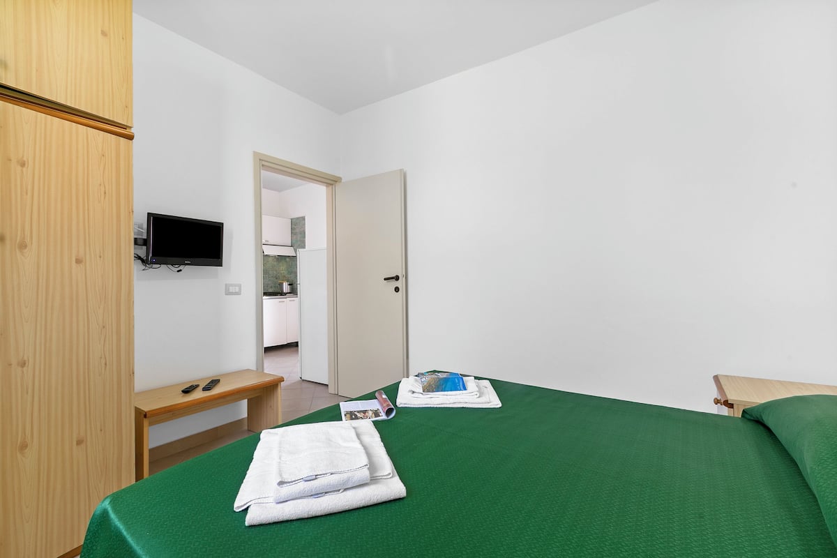 A hotel room with a green bedspread, a wooden wardrobe, a desk with a flat-screen TV, and a door leading to a bathroom.