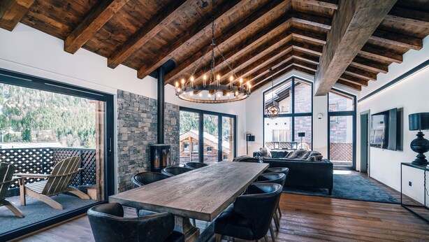 A spacious room with a large wooden table, black chairs, and a chandelier. There are large windows offering views of a mountainous landscape.