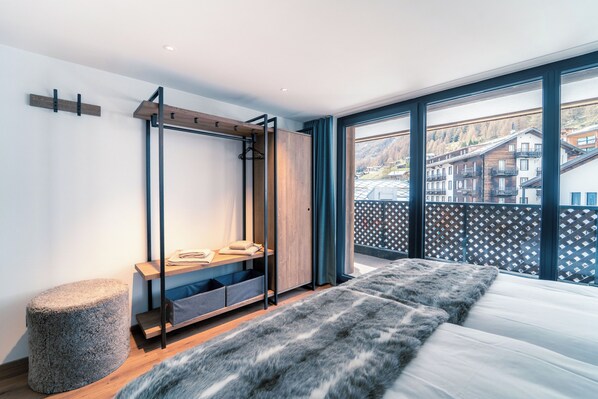 A modern bedroom with a large bed, a wooden wardrobe, a stone ottoman, and a view of a mountain town through large windows.