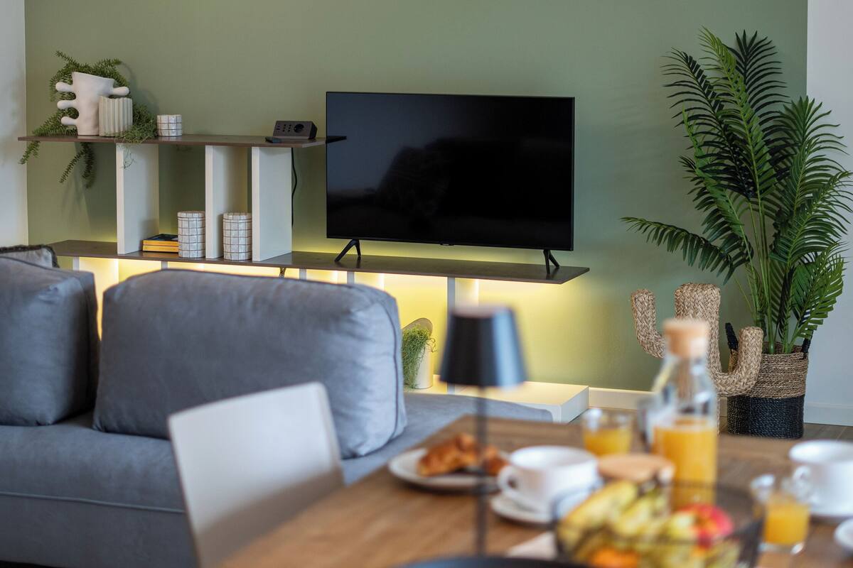 A modern living room with a flat-screen TV, a sofa, a dining table with breakfast items, and a hanging light fixture.