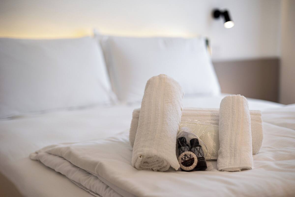 A neatly made bed with white linens and towels, a small container with toiletries, and a wall-mounted light fixture.