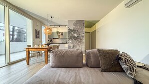 A modern living room with a sofa, pillows, a dining table, and a view of the kitchen. - CaNova (Spinea)