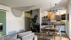 A modern living room with a grey sofa, a dining table with chairs, and a kitchen area with a hanging light fixture. - CaNova (Spinea)