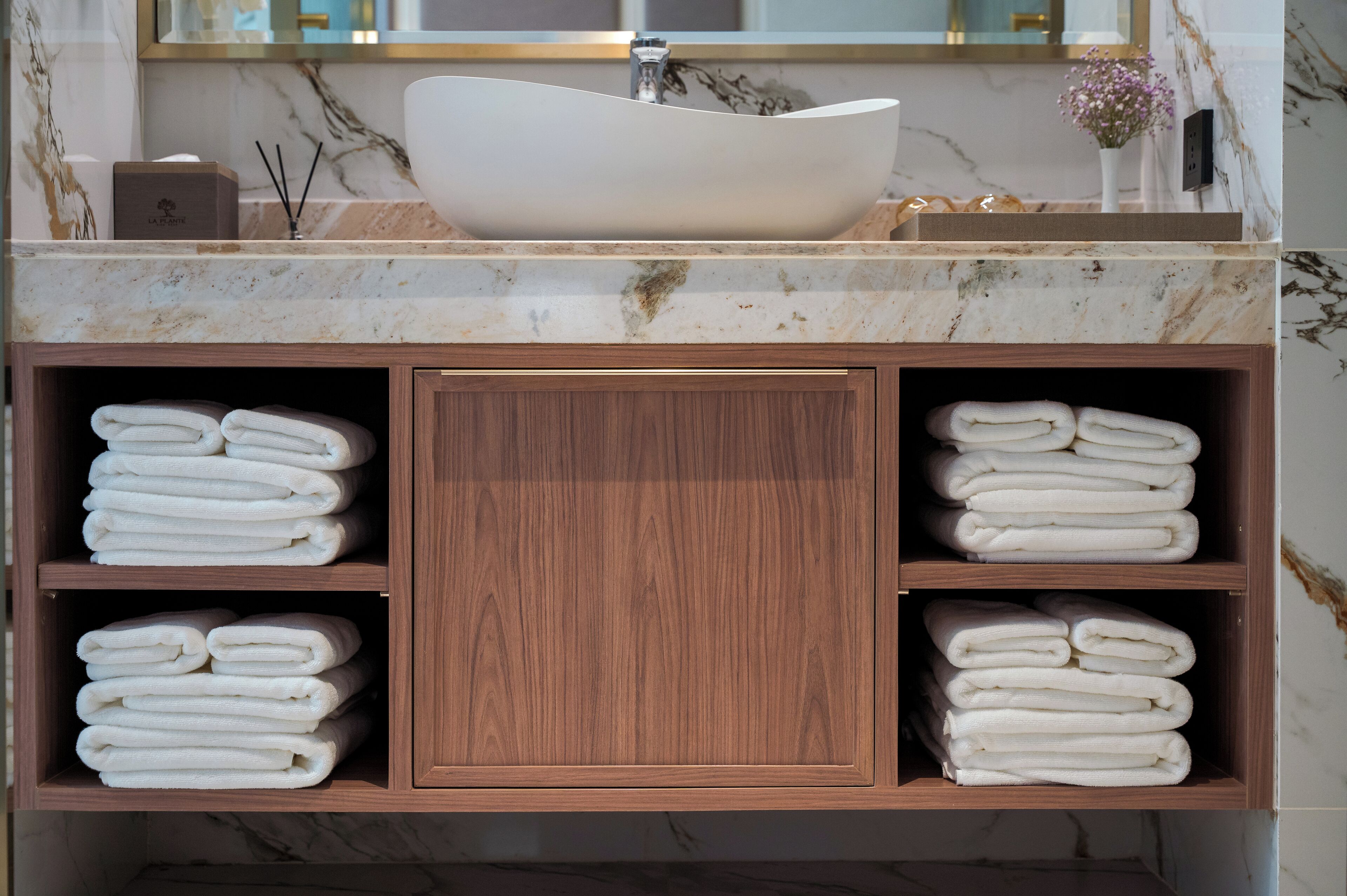 Un tocador de baño con un lavabo blanco tipo recipiente, encimera de mármol y estantes de madera con toallas dobladas.