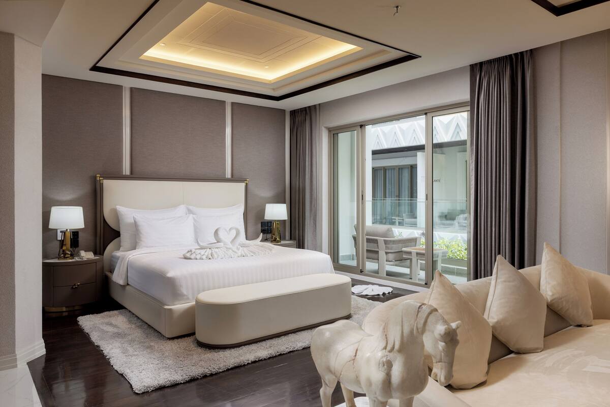 A modern hotel bedroom with a large bed, a sitting area with a sofa, and a view of the outside.