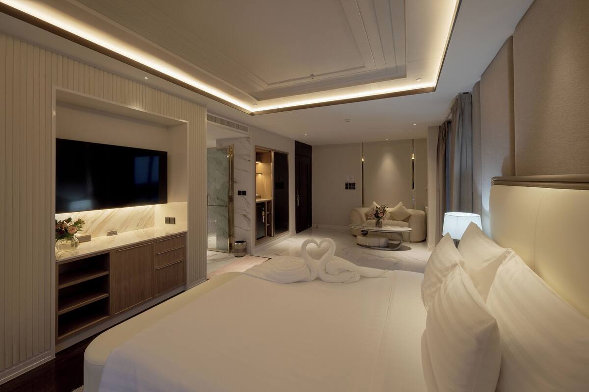 A modern hotel room with a large bed, a flat-screen TV, a marble accent wall, a seating area with a table and chairs, and a bathroom with a shower and toilet.