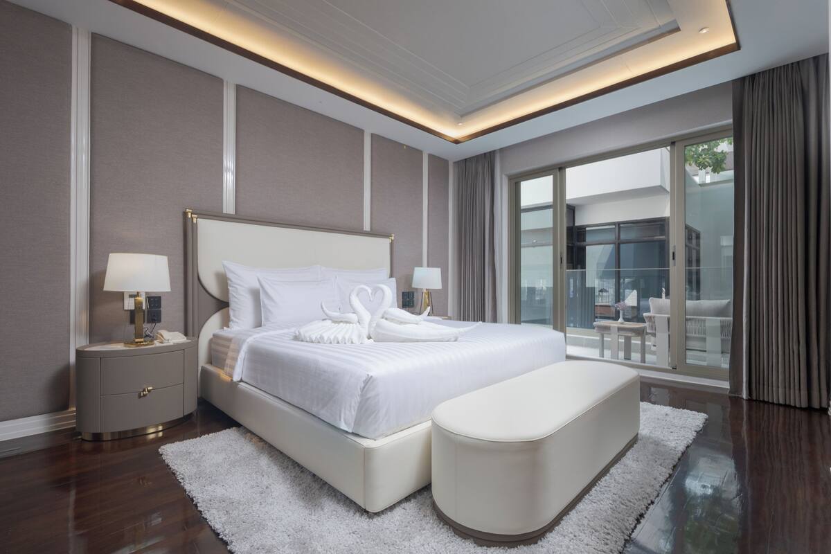 A modern hotel room with a large bed, a bedside table, a nightstand, a white upholstered bench, and a large window offering a view of the outdoors.