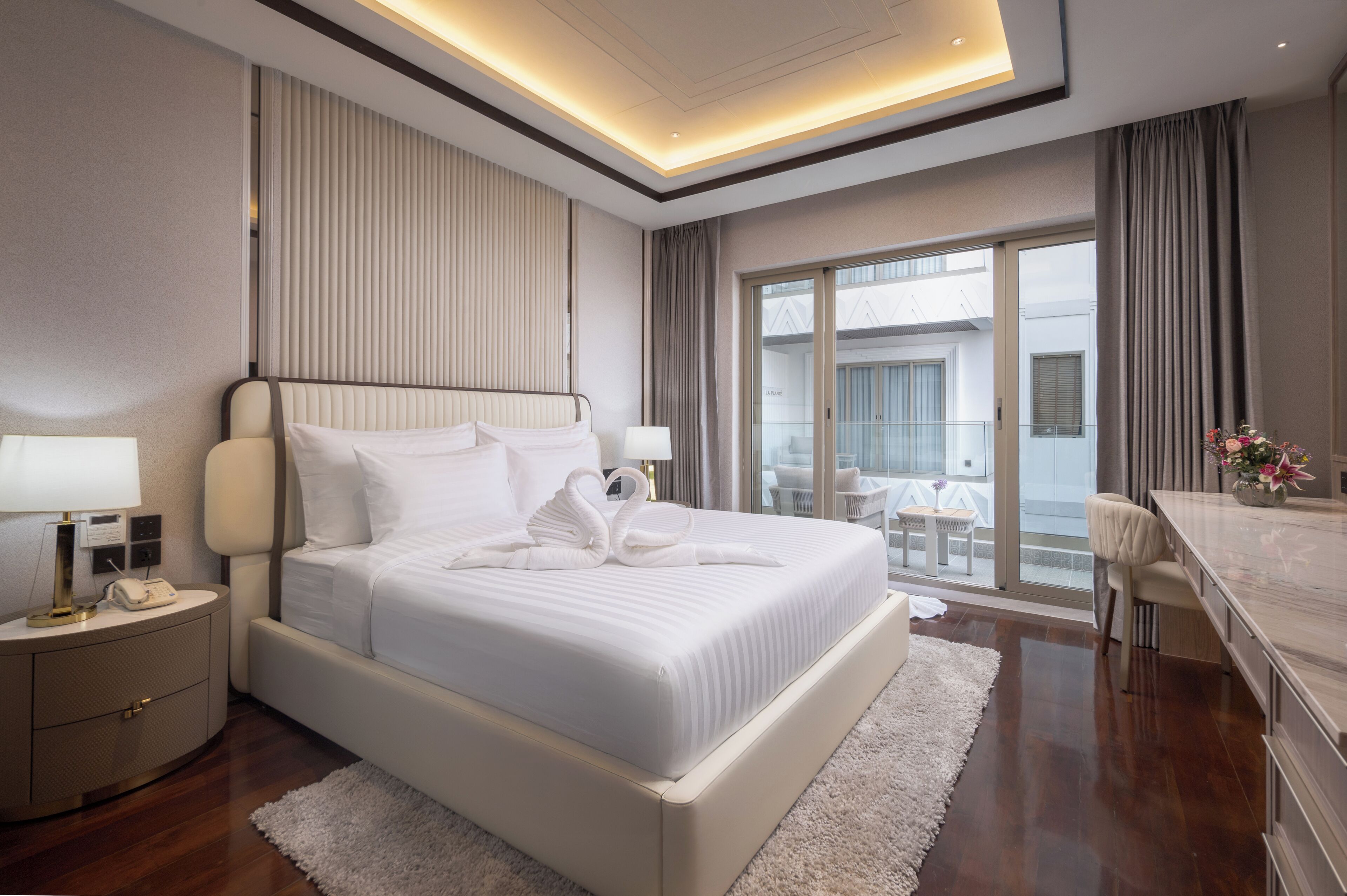 Una habitación de hotel moderna con una cama grande, mesitas de noche, un escritorio y un balcón con mobiliario exterior.
