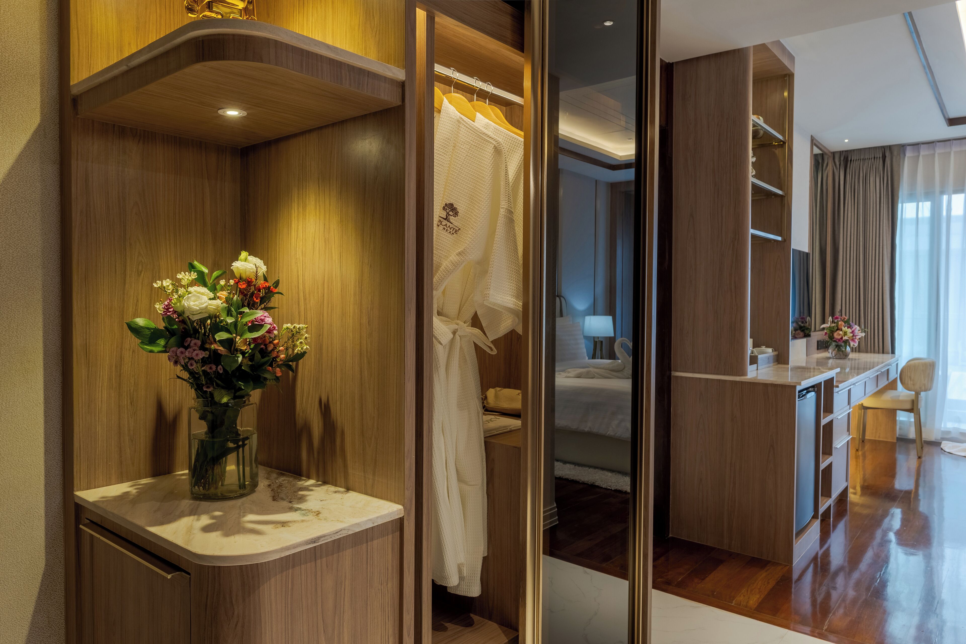 A hotel room with a wooden wardrobe, a vanity with a mirror, a bed with white bedding, a small desk with a lamp, and a vase with flowers on the vanity.