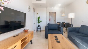 A modern living room with a flat-screen TV, wooden coffee table, blue sofa, and a dining area in the background. - Riviera Golf Apartment - Pool Views!  - Pass the Keys (Urbanización Riviera Sol)