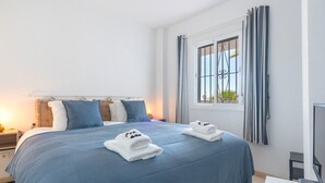 A hotel room with a large bed, a bedside table with a lamp, a TV on a stand, and a window with curtains. - Riviera Golf Apartment - Pool Views!  - Pass the Keys (Urbanización Riviera Sol)