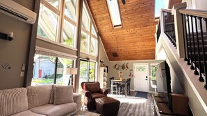 A spacious living area with a high ceiling, large windows, a staircase, and a dining area.