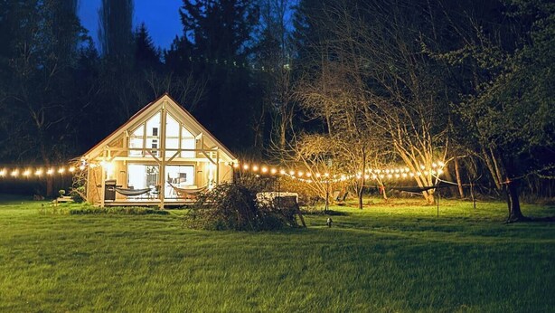 A two-story wooden cabin with a large front porch and string lights, surrounded by trees and grass.