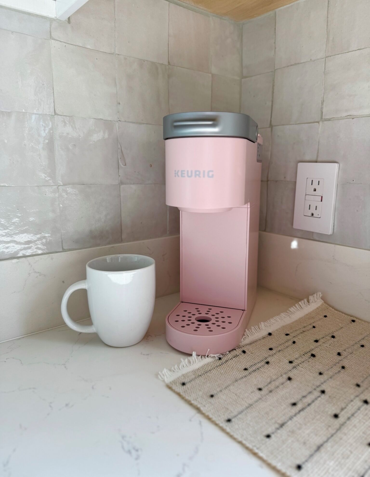 A KEURIG coffee maker in a pink color, a white mug, and a woven placemat on a tiled countertop.