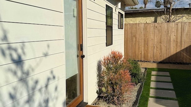 Una casa con puerta principal, un pequeño jardín con un sendero de adoquines y una valla de madera al fondo.