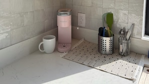Una cocina con encimera blanca, una cafetera Keurig rosa, una taza, un mueble para especias y una ventana con vistas al exterior.