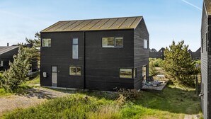 Een modern, twee verdiepingen hoog huis met een donkere gevel, meerdere ramen en een klein terras met zitplaatsen buiten.