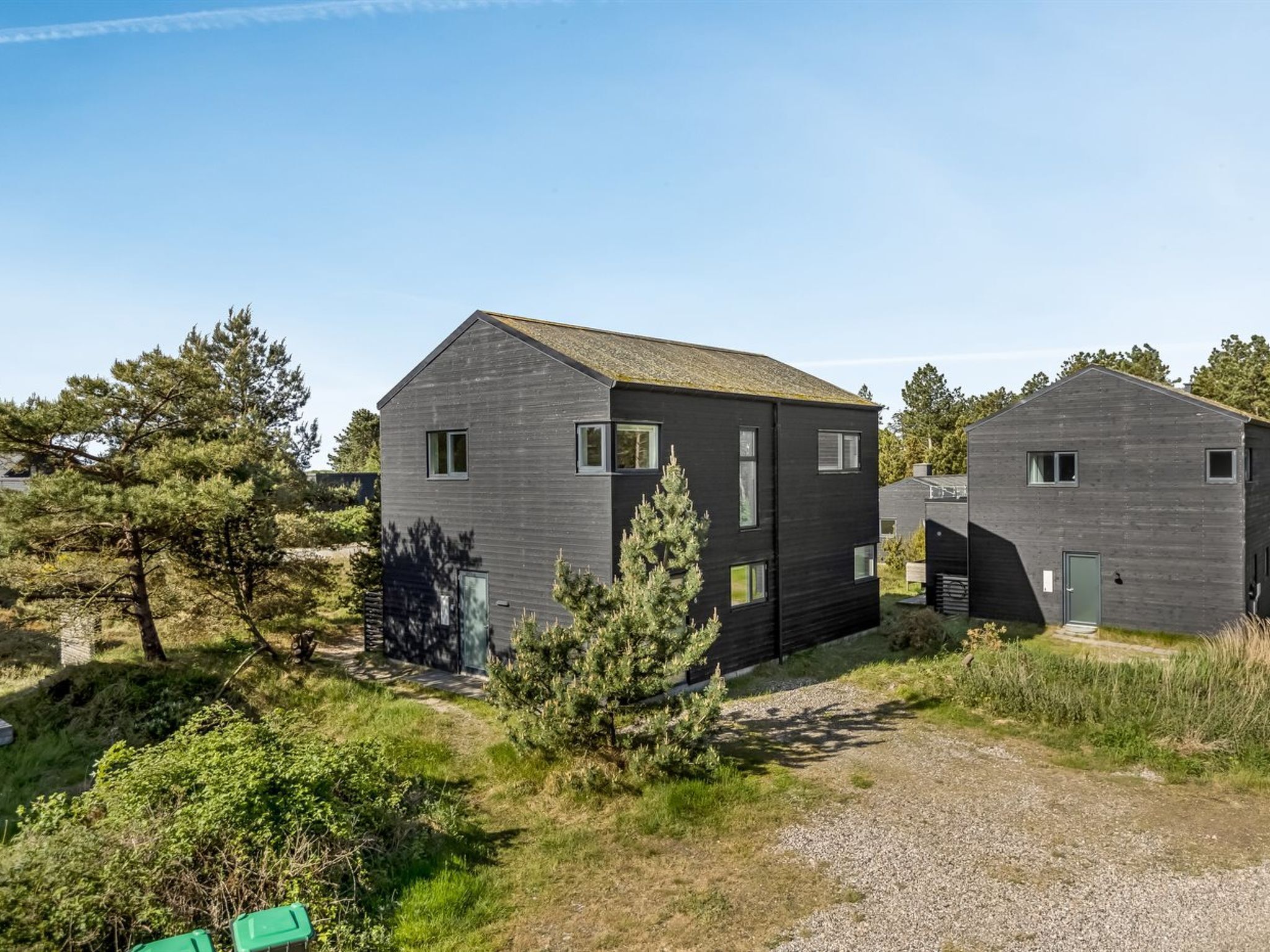 Deux cabanes modernes aux couleurs sombres, dotées d’un design simple et rectangulaire, entourées d’une végétation luxuriante et baignées par un ciel dégagé.
