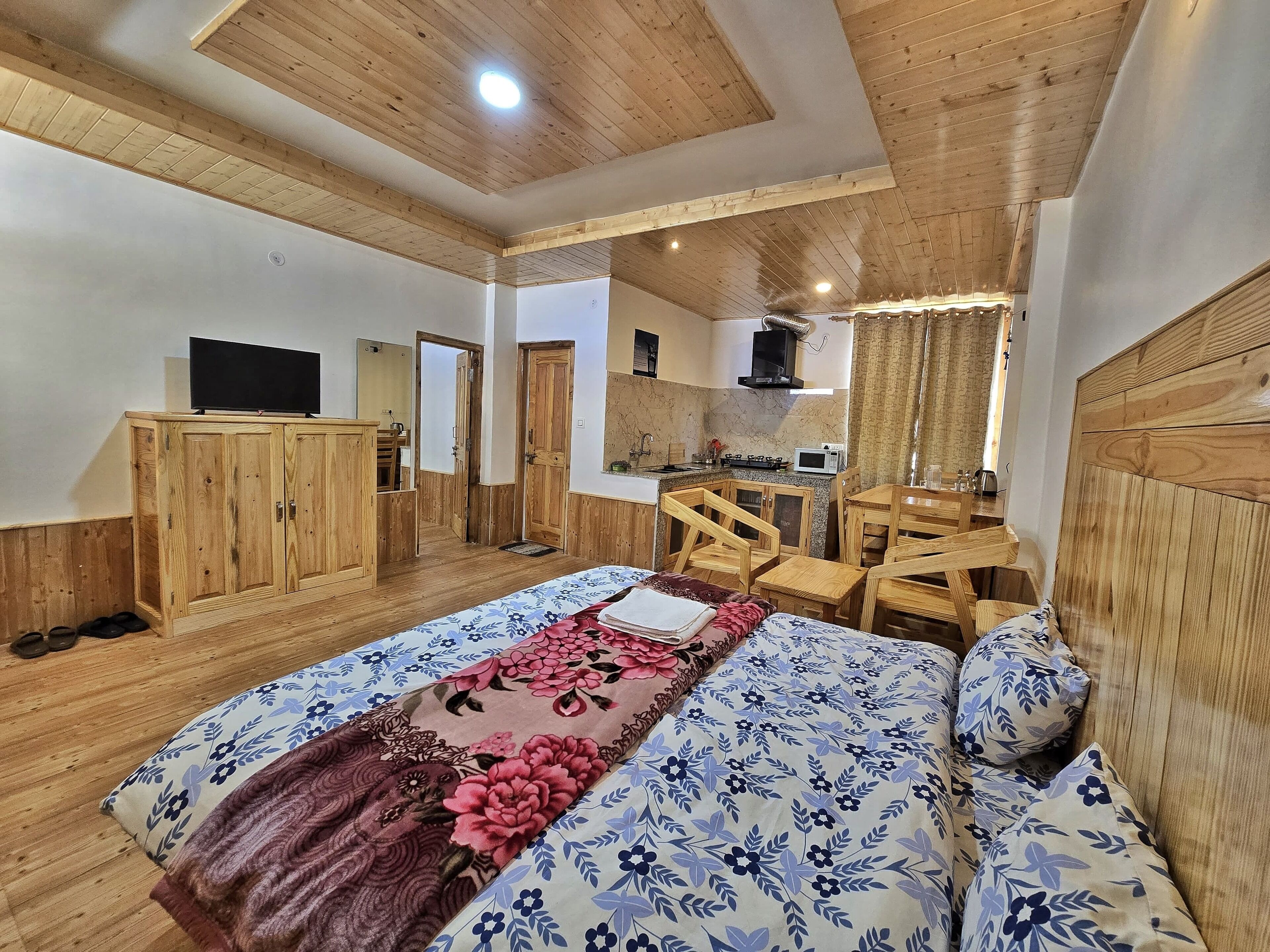 Een slaapkamer met een houten plafond, een bed met bloemenbeddelsels, een houten kledingkast en een kitchenette op de achtergrond.