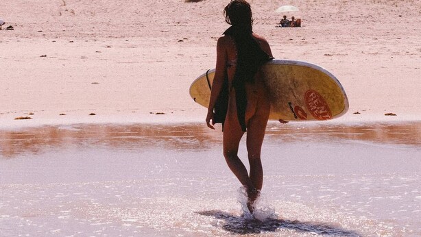A person carrying a surfboard walking on a sandy beach with dunes in the background. - Guesthouse Beach Break (Peniche)