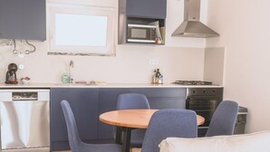 A compact kitchen with a microwave, stove, and sink. There is a dining table with chairs and a sofa with a floral pillow. - Guesthouse Beach Break (Peniche)