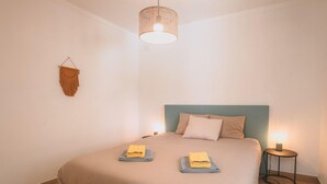 A neatly made bed with two folded towels, a hanging light fixture, and a small bedside table with a lamp. - Guesthouse Beach Break (Peniche)