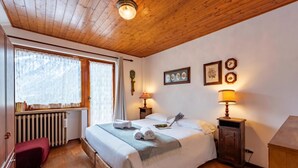 Une chambre avec un plafond en bois, un lit recouvert de draps blancs, deux lampes de chevet, un radiateur et une fenêtre avec des rideaux.