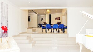 A spacious living area with a white grand piano, a dining area with blue chairs, and a staircase leading to another room.