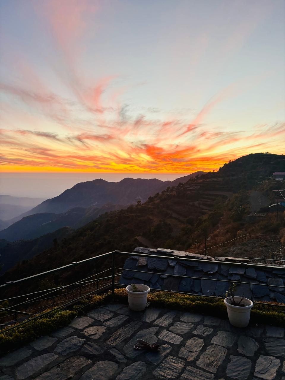 A stone-paved terrace with potted plants, overlooking a mountainous landscape during sunset.