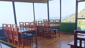 A dining area with wooden chairs and tables, large windows with a view of hills, and a wooden ceiling. - Deodar Inn resort (Kalsi)