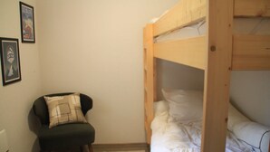 Une petite pièce avec un lit superposé, une chaise avec un coussin et deux tableaux encadrés accrochés au mur.