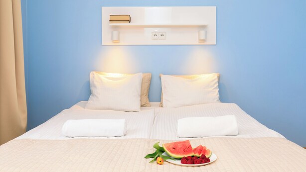 Un lit bien fait, avec du linge de lit blanc, une table de chevet sur laquelle repose un livre, et une assiette de fruits posée sur le lit.