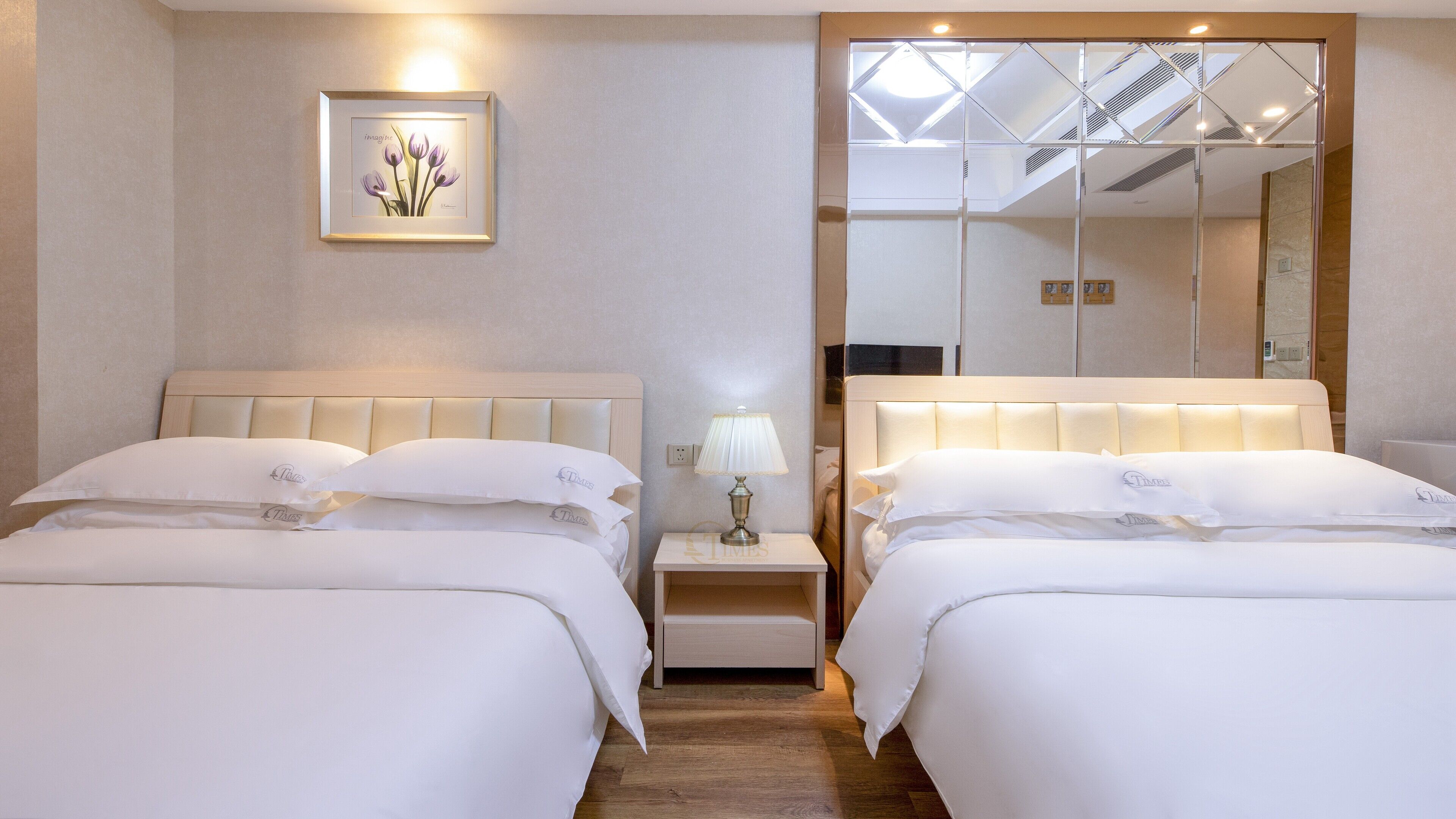 A hotel room with two beds, a bedside table with a lamp, a framed picture on the wall, and a large mirror.