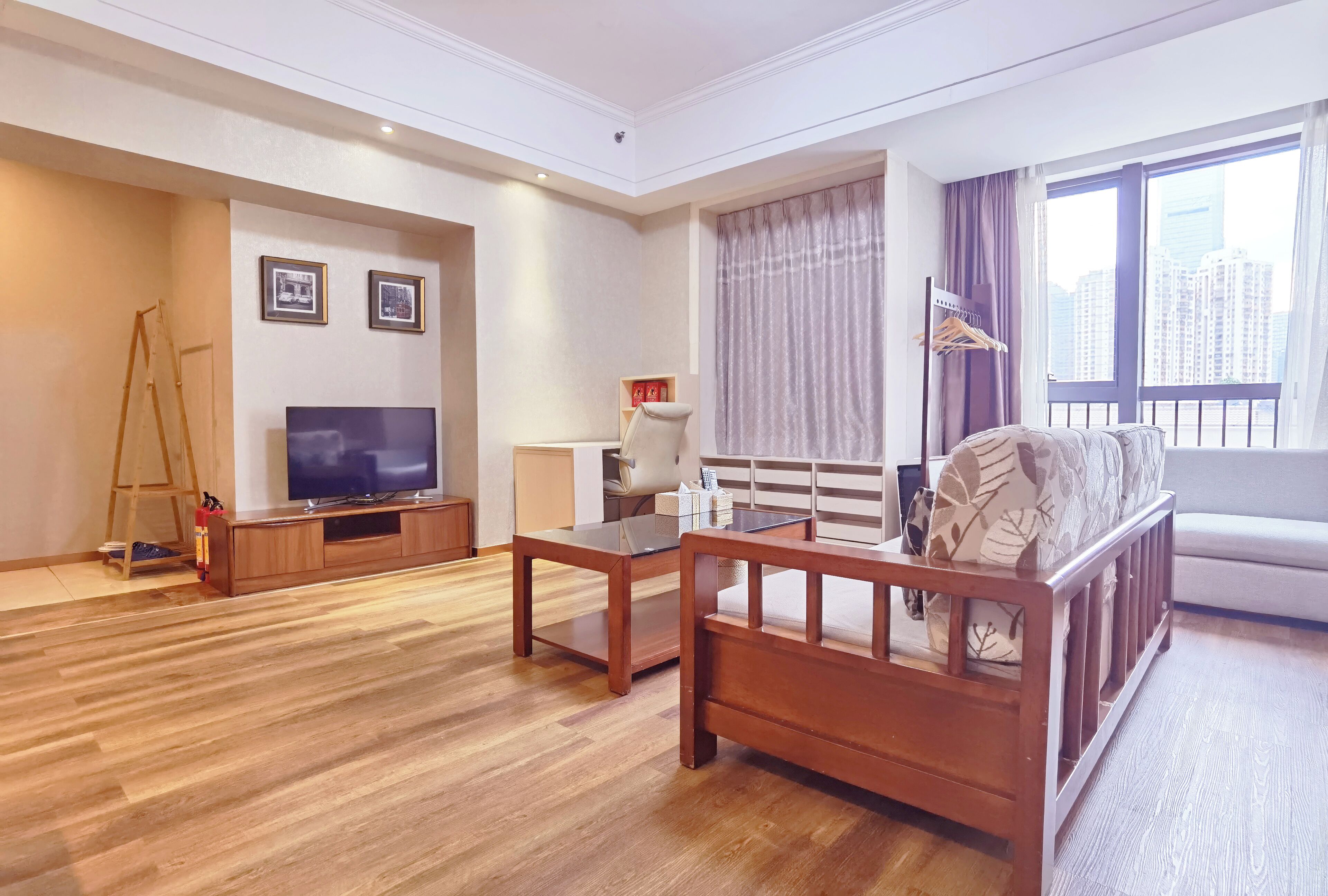 A modern living room with a flat-screen TV, wooden furniture, a sofa with patterned cushions, a window with curtains, and a city view.