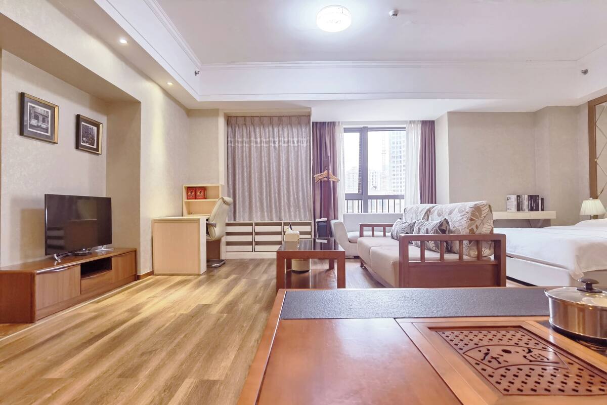 Habitación de hotel con piso de madera, televisor, sofá, cama, una mesita y ventana con cortinas.