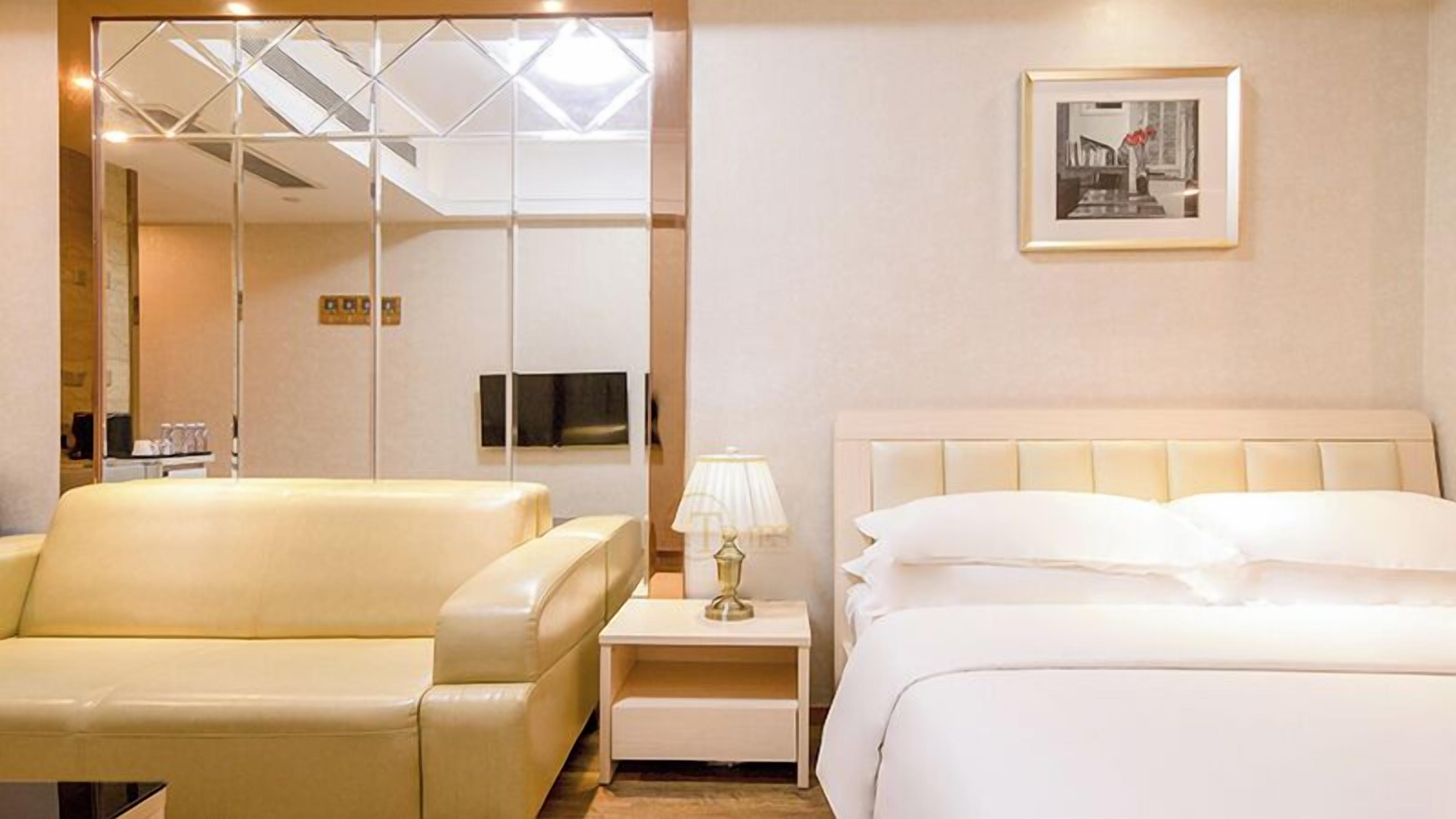 A hotel room with a bed, a sofa, a nightstand with a lamp, a TV, and a framed picture on the wall.
