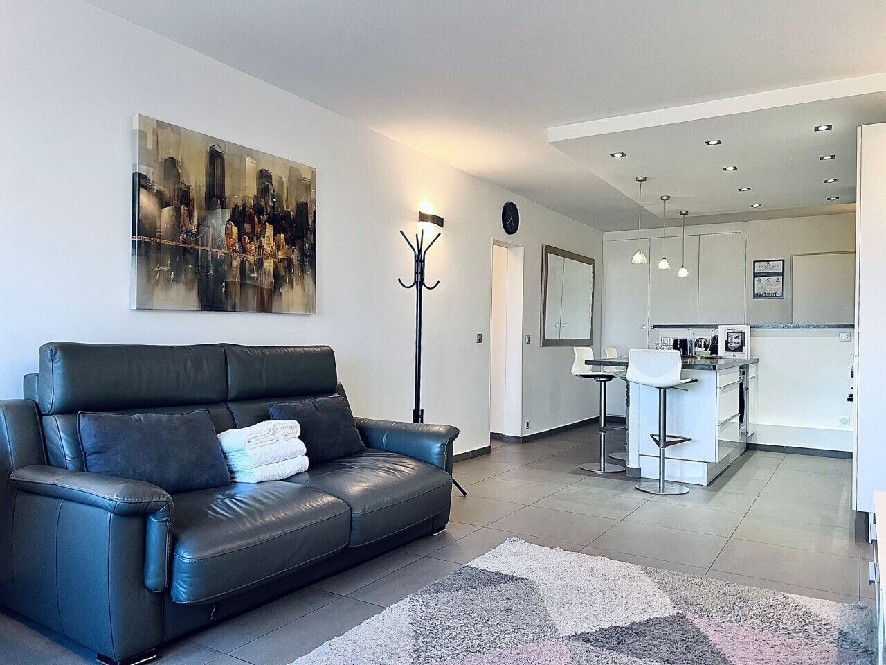 A modern living room with a black leather sofa, a large abstract painting, a floor lamp, and a kitchen area with white cabinets and a bar stool.