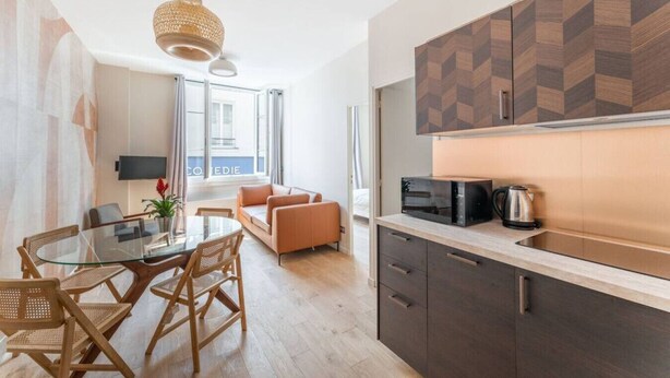 A modern kitchen with dark cabinets, a microwave, and a kettle. There is a dining area with a glass table and wicker chairs. - Charming home in heart of PARIS!Opera/Eurostar 4pers (Paris)