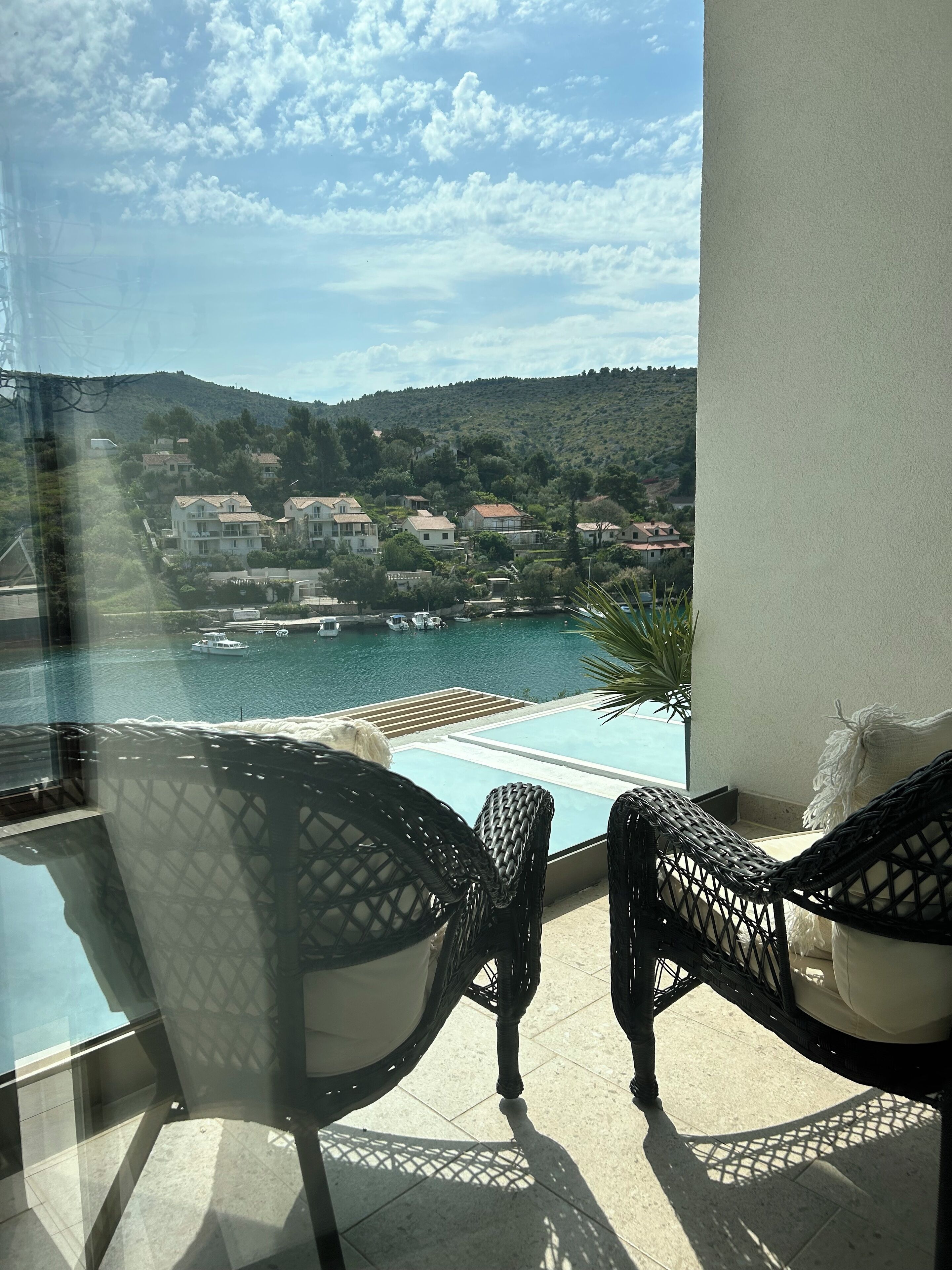 A balcony with two wicker chairs, a glass table, and a view of a coastal town with houses and boats.