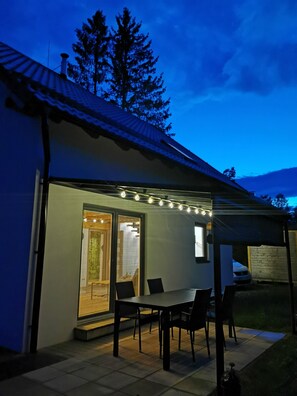 A covered patio area with a table and chairs, a window with blinds, and string lights.