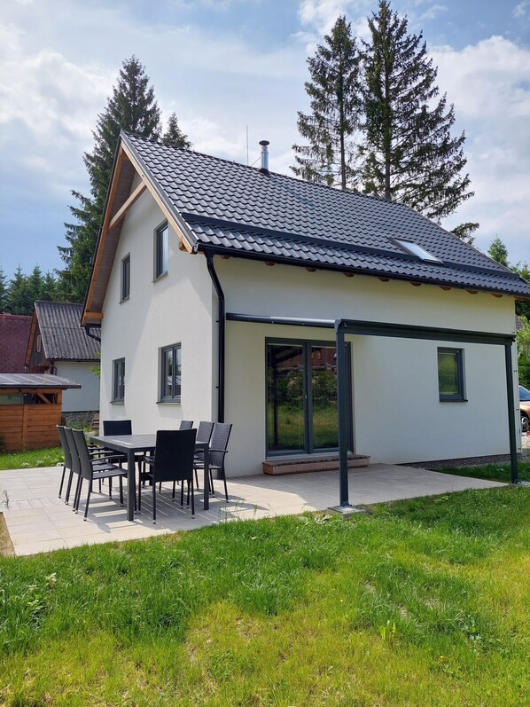 A modern two-story house with a sloped roof, large sliding glass doors, and a patio with outdoor furniture.