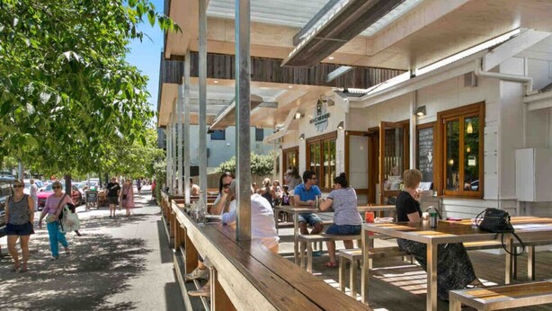 An outdoor café with wooden tables and benches, a clear sky, and a few people dining. - Private Barwon Heads Cottage | Walk to River (Barwon Heads)