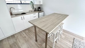 A modern kitchen with white cabinets, a wooden table, and a window with blinds. - Private Barwon Heads Cottage | Walk to River (Barwon Heads)