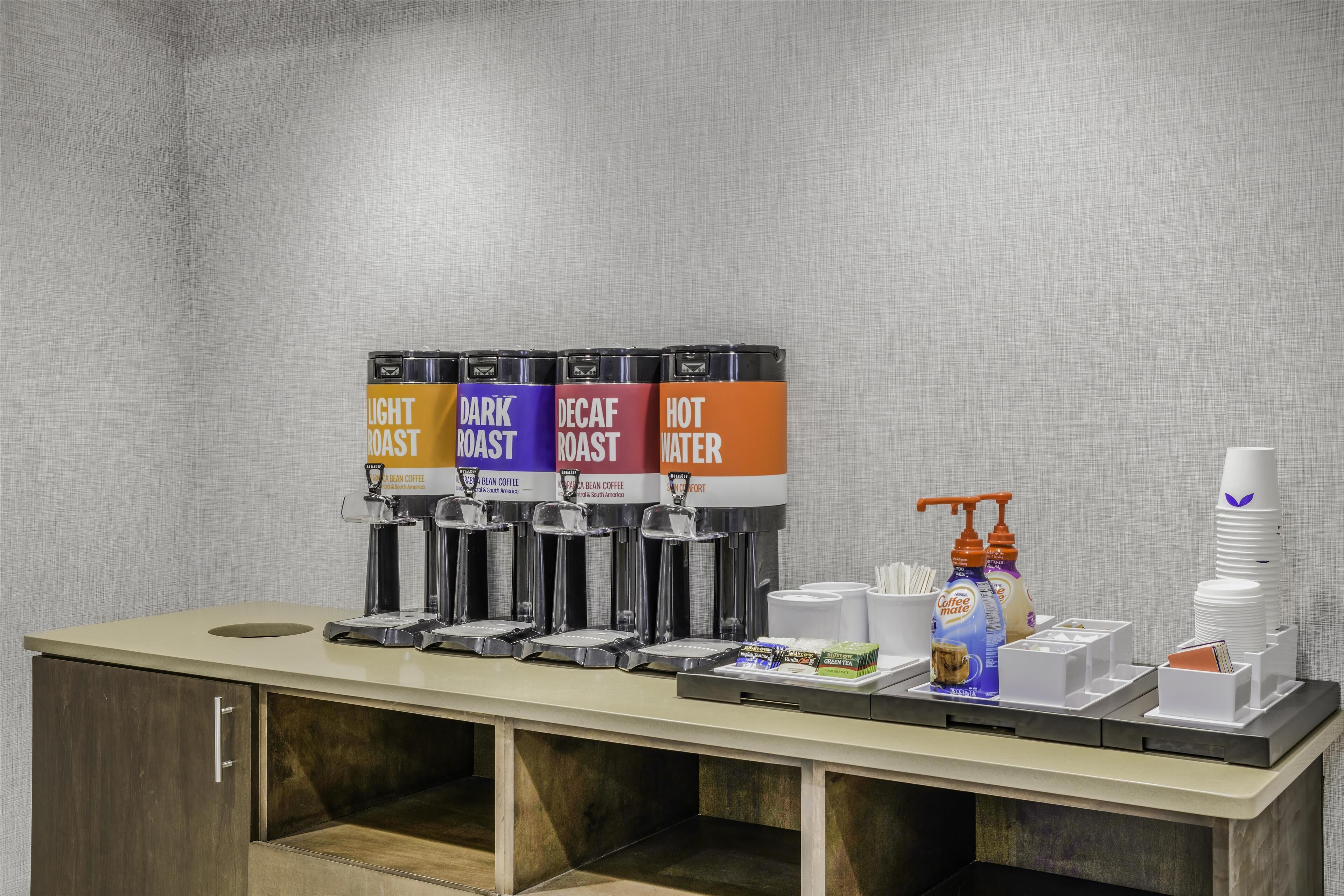 A coffee station with four dispensers labeled 'Light Roast,' 'Dark Roast,' 'Decaf Roast,' and 'Hot Water.' There are also disposable cups, a coffee maker, and a bottle of hand sanitizer.