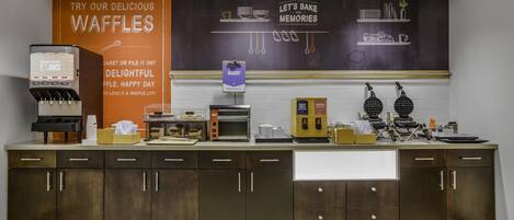 A waffle station with a sign promoting waffles, a juice dispenser, and a bakery section.