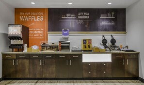A waffle station with a sign promoting waffles, a juice dispenser, and a bakery section.